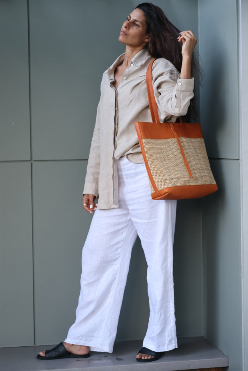 Handmade Venice rattan tote bag featuring camel-toned leather straps and natural rattan body. Unique strap closure for a chic and secure look.