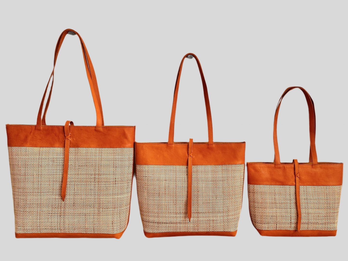 A view of all 3 sizes of the handmade Venice rattan tote bag featuring camel-toned leather straps and natural rattan body. Unique strap closure for a chic and secure look.